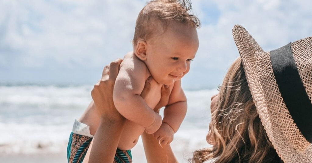 main image of things to do with a newborn in summer: mom holding baby by the ocean
