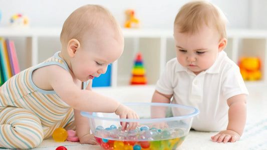 A child's hand uses a hammer to drive nails into small wooden blocks on a colorful, paint-splattered table. A packet of screws and toy parts lies nearby.