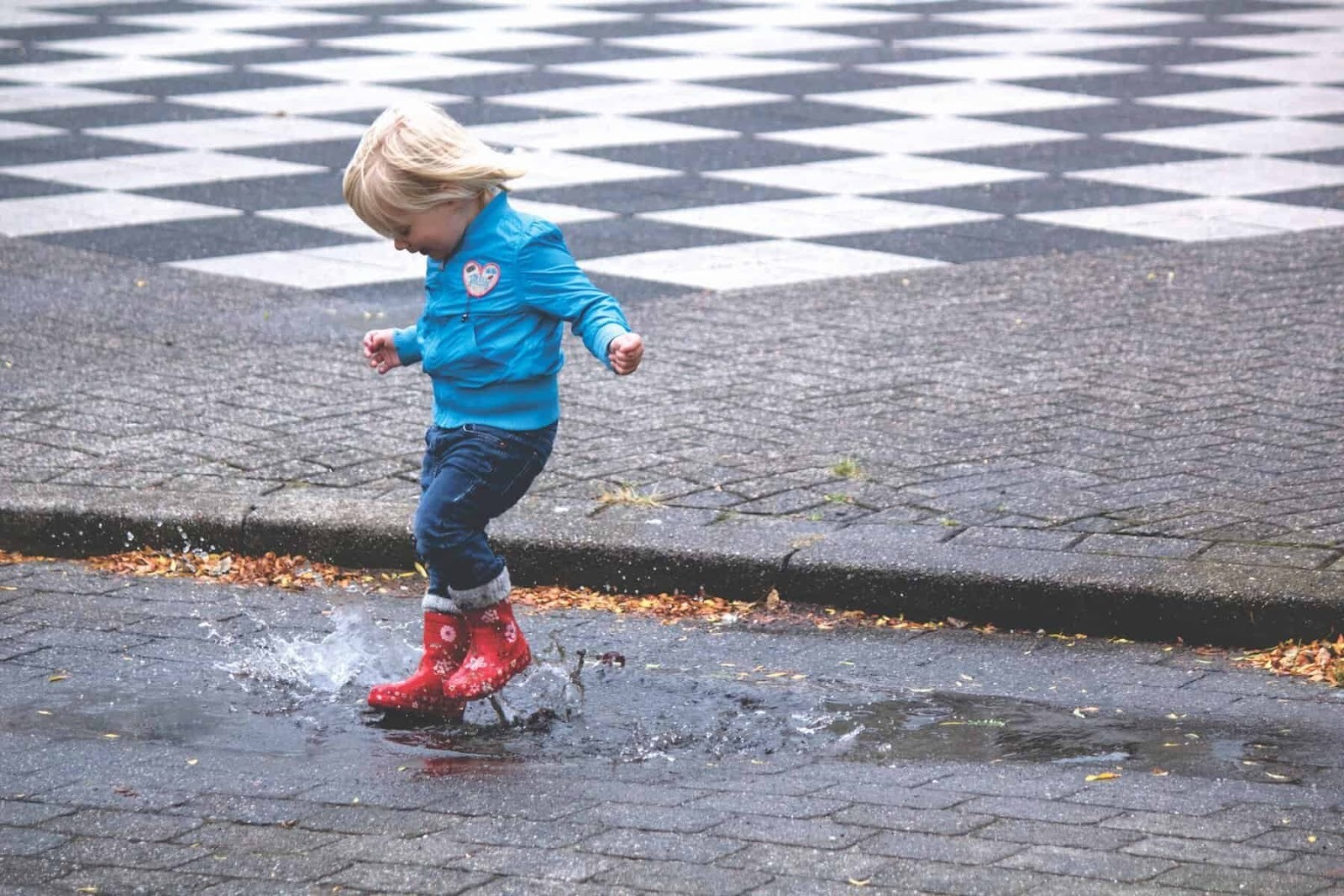 a young child jumping in puddles wearing red rubber boots, jeans and a blue jacket
