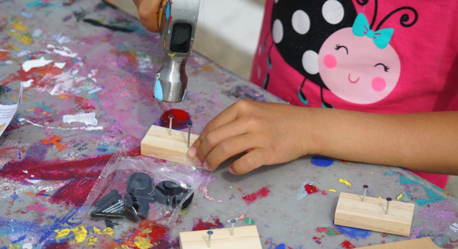 A child's hand uses a hammer to drive nails into small wooden blocks on a colorful, paint-splattered table. A packet of screws and toy parts lies nearby.